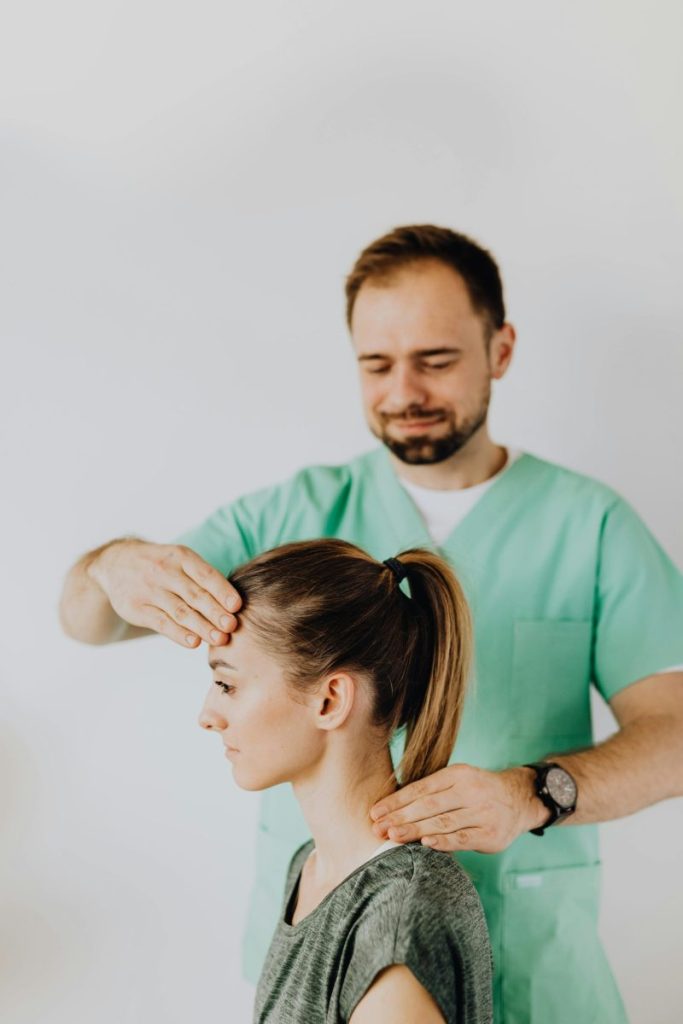 Positive bearded doctor in uniform doing rehabilitation neck massage on female patient after injury against gray background