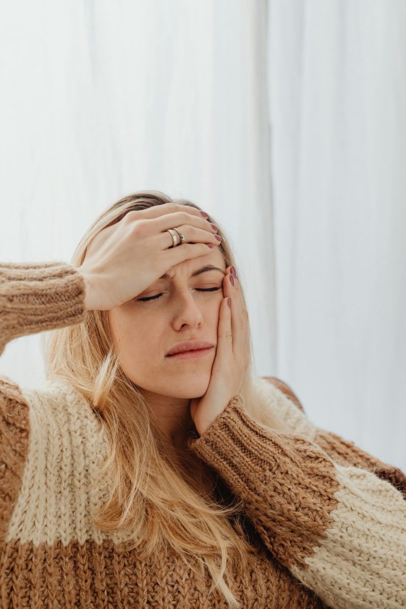 Woman with closed eyes holding head, wearing a knitted sweater, possibly experiencing discomfort.